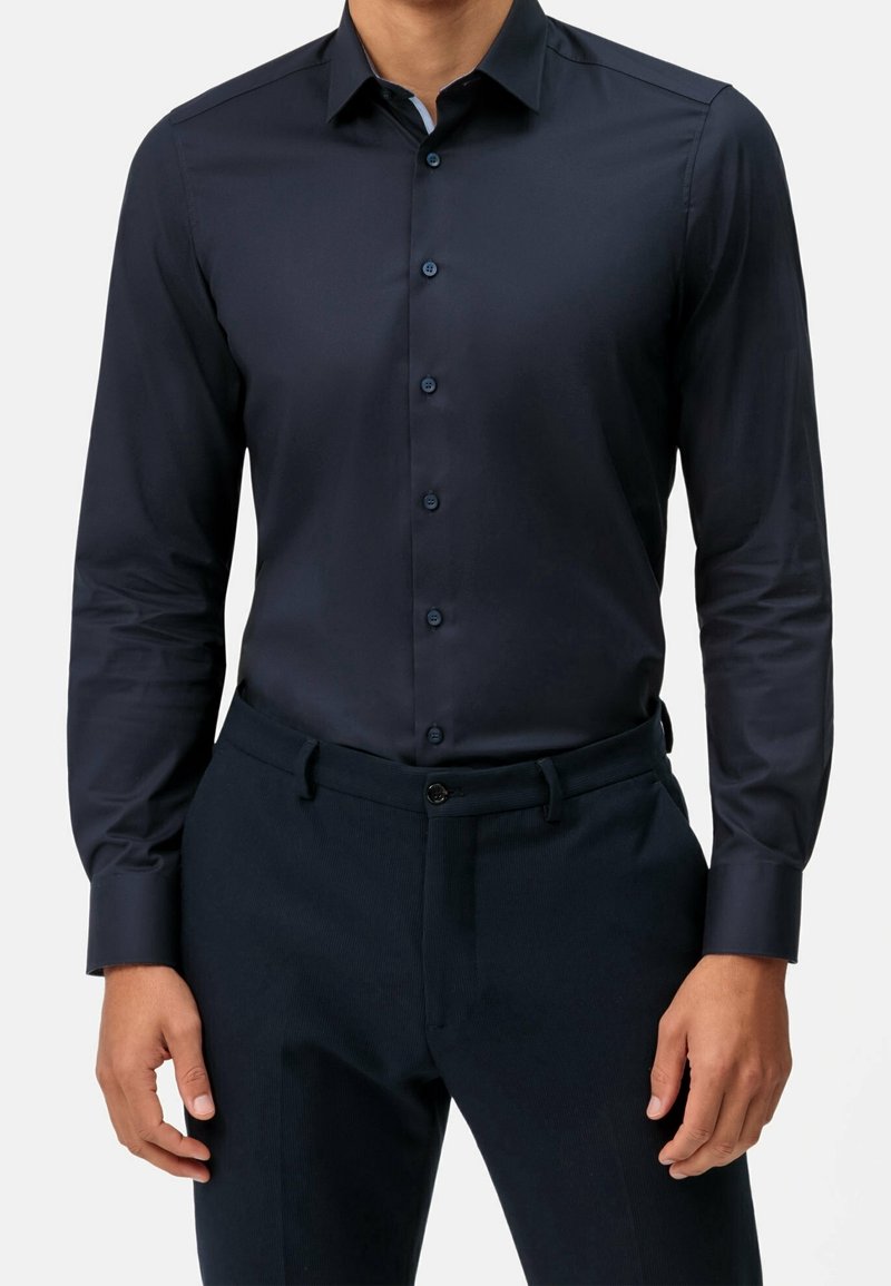 Homme portant une chemise ajustée bleu marine foncé à manches longues et boutonnée, rentrée dans un pantalon bleu marine assorti avec une ceinture noire à bouton.