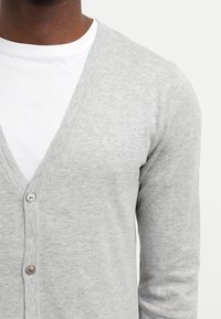 Cárdigan gris claro con cuello en V, con un frente abotonado y un logo bordado simple. Llevado sobre una camiseta blanca. Textura suave.