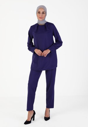 Purple tunic with long sleeves and a rounded neckline; paired with matching trousers. The top features a subtle chain detail at the collar.