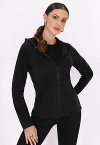 Schwarze Zip-Hoodie aus weichem Material, mit Kapuze, langen Ärmeln und figurbetontem Schnitt, kombiniert mit einem schwarzen Oberteil und schmalen Hosen.