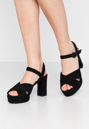 Jambes portant des sandales à talons compensés en daim noir avec des brides croisées et une fermeture à boucle à la cheville sur un sol gris clair.