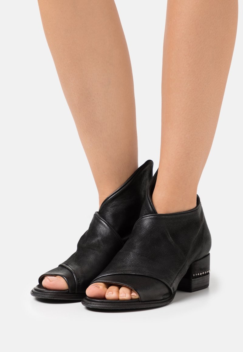 Bottines en cuir noir à bout ouvert avec talons blocs bas portées pieds nus sur un fond clair uni.