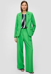 Costume sur mesure vert avec un pantalon à jambes larges et une veste ajustée. En dessous, un chemisier à motifs présente des rayures bleu marine, blanches et vertes.
