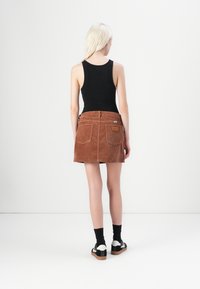 Brown corduroy mini skirt with front and back pockets, paired with a black tank top and black socks. Model facing away, showing skirt texture.