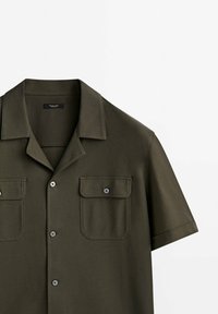 Chemise vert olive à manches courtes avec col camp, fermeture boutonnée devant et deux poches poitrine avec rabats boutonnés.
