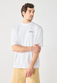 Hvit bomull t-skjorte med korte ermer, med teksten "Soar into Tranquility" i svart på venstre bryst. Enkel design, ingen mønstre.