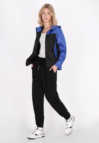Svart och blå vindjacka med dragkedja framtill, elastiska ärmslut och huva, kombinerad med svarta sweatpants och vita sneakers.