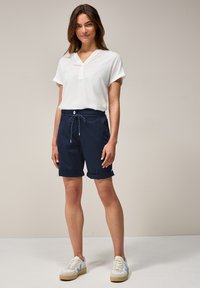 Marineblauwe shorts met verstelbaar trekkoord, zijzakken, opgerolde manchetten, gecombineerd met een witte blouse en casual sneakers.