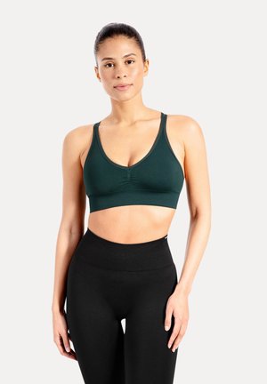 Mujer de pie mirando hacia adelante, con un sujetador deportivo verde oscuro y leggings negros de talle alto sobre un fondo claro y liso.