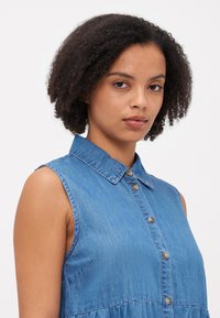 Τζιν φόρεμα, ανοιχτό γαλάζιο, χωρίς μανίκια με γιακά. Διαθέτει κουμπί και μαζεμένη μέση. Απαλή υφή με διακριτική γυαλάδα.