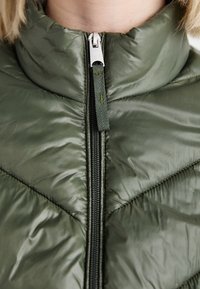 Grønn, quiltet puffervest med høy krage og glidelås. Den har en blank tekstur og skrå sømmer.