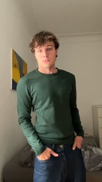 Jovem com cabelo castanho a usar um suéter verde e jeans azuis está de pé com as mãos nos bolsos numa sala com arte de parede abstrata e uma cama.