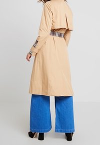 Donna che indossa un trench beige con cintura e polsini a quadri, abbinato a jeans a gamba larga blu e scarpe con tacco nere, di spalle.