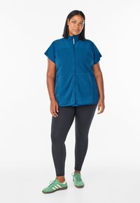 Turkusowa kamizelka polarowa z krótkimi rękawami, pełnym zamkiem z przodu, kieszeniami z przodu, noszona na czarnych legginsach i zielonych trampkach z białymi akcentami.