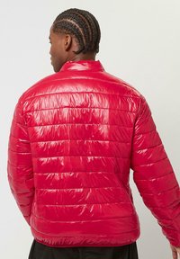 Rote, puffige Jacke mit glänzender Oberfläche, gestepptem Design, hohem Kragen und elastischen Bündchen. Leicht und warm mit horizontalen Nähten.
