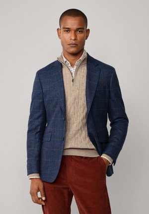 Marineblauer karierter Blazer über einem beigen Kabelstrickpullover, kombiniert mit rostfarbenen Cordhosen. Einkragenhemd mit sichtbarem Karomuster.