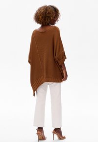 Brązowy oversize'owy sweter z dzianiny o szerokim dekolcie i podwiniętych rękawach. Ma strukturalny splot i dłuższy tylny brzeg. W zestawieniu z białymi spodniami.