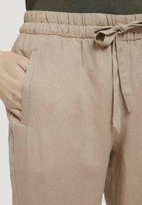 Pantalons en lin beige avec une taille élastique, cordon de serrage ajustable et poches latérales. Le tissu semble léger avec une texture subtile.