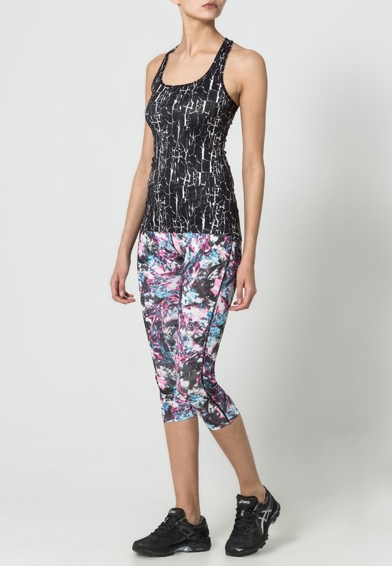 Femme portant un haut sans manches noir à motifs, des leggings capri abstraits multicolores et des chaussures de sport noires, debout devant un fond uni.