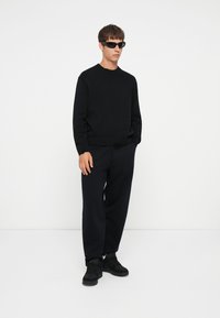 Schwarzer Strickpullover mit Rundhalsausschnitt, kombiniert mit locker sitzenden schwarzen Jogginghosen und schwarzen Sneakers. Elegante schwarze Sonnenbrille rundet den Look ab.