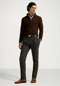 Suéter de punto marrón con cuello de media cremallera, combinado con pantalones de tela gris con pliegues y zapatos de cuero marrón. Los accesorios incluyen gafas transparentes y una camisa azul.