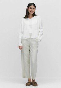 Witte cardigan met open voorkant, gecombineerd met een witte top en lichtbeige, wijde broek. Bruine moccasin-achtige slippers maken de look compleet.