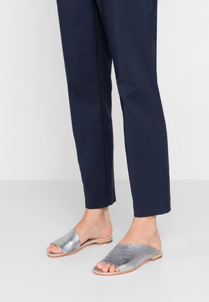 Hopeakylkiset metalliset liukusandaalit, joissa on sileä pinta, avoin varvasmuotoilu ja litteä pohja, yhdistettynä tummansinihin housuihin kontrastiksi.