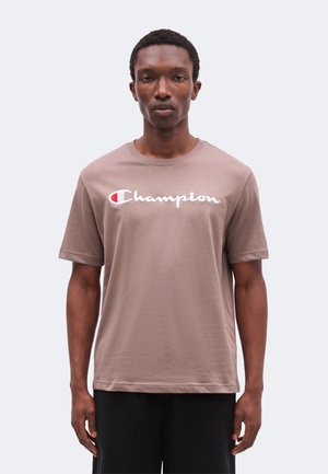Jeune homme debout face à nous, portant un t-shirt Champion marron clair avec un texte de logo blanc et rouge sur un fond uni.