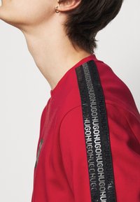 Felpa rossa con collo tondo e banda laterale nera con logo del marchio ripetuto. Il tessuto è liscio con una leggera lucentezza.