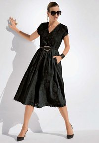 Schwarzes Kleid mit V-Ausschnitt, floraler Stickerei, kurzen Ärmeln, einer taillierten Taille mit einem Gürtel und einem A-Linien-Rock. Kombiniert mit spitzen schwarzen Absätzen.