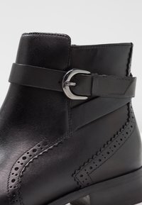 Svart läderankelsko med slät yta, utrustad med en sidoremm och spänne, invecklade perforerade detaljer och en klossklack.