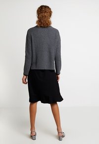 Pull en maille côtelée grise à manches longues, coupe courte, avec un ourlet arrondi, porté sur une jupe évasée noire, associé à des chaussures à motifs.