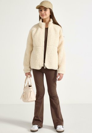 Roomkleurig pluizig jasje met een hoge kraag, gecombineerd met een bruin aansluitend outfit, beige pet en lichte handtas. Inclusief witte sneakers.