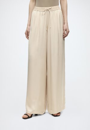 Løse beige vidde bukser med elastik i taljen og snøre, båret med sorte sandaler med åben tå, vist fra talje til fødder.