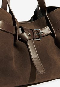 Pruun suede käekott, millel on silmapaistev nahast rihm koos hõbedase klambriga ja tekstureeritud pind. Käepidemed on elegantsed ja vastupidavad.