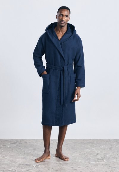 Άνδρας που στέκεται ξυπόλυτος σε πλακάκια φορώντας μια μπλε-σκούρα μπαντάνα με κουκούλα, μπροστινές τσέπες και δεμένη ζώνη.