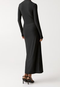 Robe noire à manches longues en matière lisse, présentant une coupe ajustée avec une taille légèrement froncée et une longueur maxi, portée avec des talons à brides.