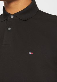 Polo noir avec un col, deux boutons et un tissu texturé. Présente un petit logo rouge, blanc et bleu sur la poitrine gauche.