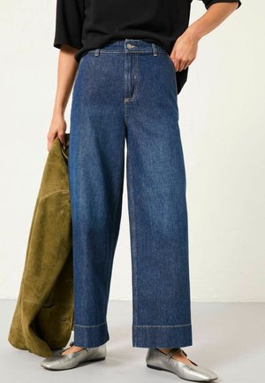 Wide-leg denim jeans in dark blue, featuring a high waist, front button closure, and contrast stitching. Paired with a green suede jacket.