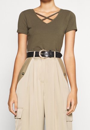 Olivgrünes V-Ausschnitt-T-Shirt mit gekreuzten Trägern, kombiniert mit hoch taillierten beigen Hosen mit Seitentaschen und einem verstellbaren schwarzen Gürtel.