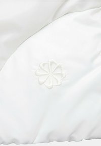 Vit vadderad tyg med en subtil lyster, prydd med en broderad blommönster i matchande färg nära kanten. Slät yta.