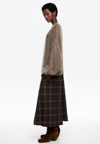 Žena stojaca bokom, oblečená v voľnom hnedom chlpatom svetri, s dlhou tmavou kockovanou sukňou a hnedými semišovými členkovými čižmami na bielom pozadí.