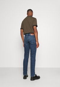 Jeans de denim azul oscuro con corte slim, que cuentan con una parche de cuero en la parte trasera de la cintura, y una camisa verde oliva lisa con acentos negros.