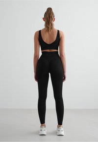 Juoda sportinė liemenėlė su giliais V formos nugara, derinama su aukštu liemeniu juodomis leggins. Modelis vilki baltus sportinius batelius. Lygus audinys, prigludęs dizainas.
