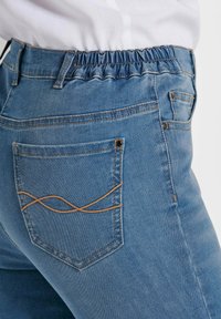 Denim jeans met een elastische tailleband, lichtblauwe kleur en een achterzak met een decoratief gestikt patroon in bruin.