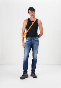 Čierny bezrukávový top, vyblednuté modré džínsy s opotrebovanými detailami, čierne tenisky a oranžová crossbody taška. Jednoduché pozadie.