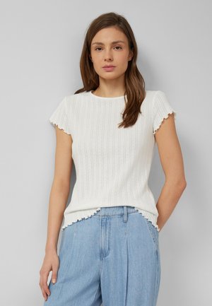 Femme aux longs cheveux bruns portant un haut blanc côtelé à manches courtes et un jean taille haute bleu clair sur un fond gris uni.