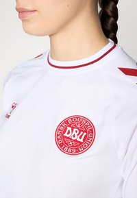 Hvid trøje med røde accenter. Har en rund hals, logo på venstre side og røde striber på skuldrene. Glat, atletisk stof.