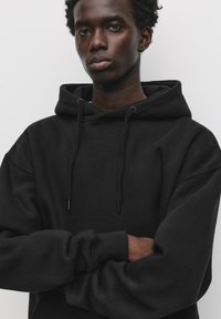 Black hoodie with a spacious fit, featuring a drawstring hood and ribbed cuffs. Made from soft, textured fabric.