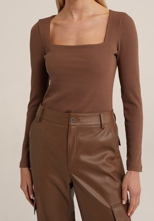Kobieta w brązowym, prążkowanym topie z kwadratowym dekoltem i długimi rękawami, włożonym w brązowe skórzane spodnie z wysokim stanem, zapinane na guziki i z kieszeniami.